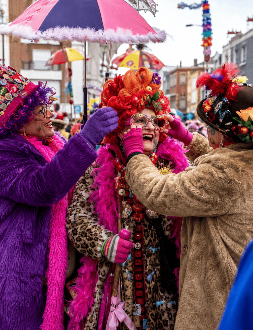 Des carnavaleux portent des perruques extravagantes, lunettes géantes, chapeaux décorés, boas, gants, maquillage et manteaux en fausse fourrure dans une rue festive inspirée du Carnaval de Dunkerque.