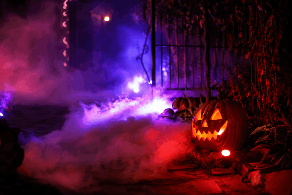Scène Halloween extérieure en gros plan avec brouillard dense au ras du sol sur une allée en pierre, citrouille sculptée illuminée, éclairages LED violets et rouges, fumée théâtrale et silhouette de portail en fer forgé dans une ambiance maison hantée.
