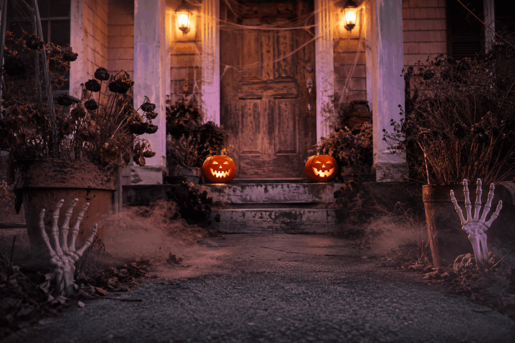 Entrée de maison décorée pour Halloween avec porte en bois vieilli, citrouilles lumineuses, toiles d’araignée, roses noires fanées, mains de squelette surgissant du sol et brume violette au crépuscule.