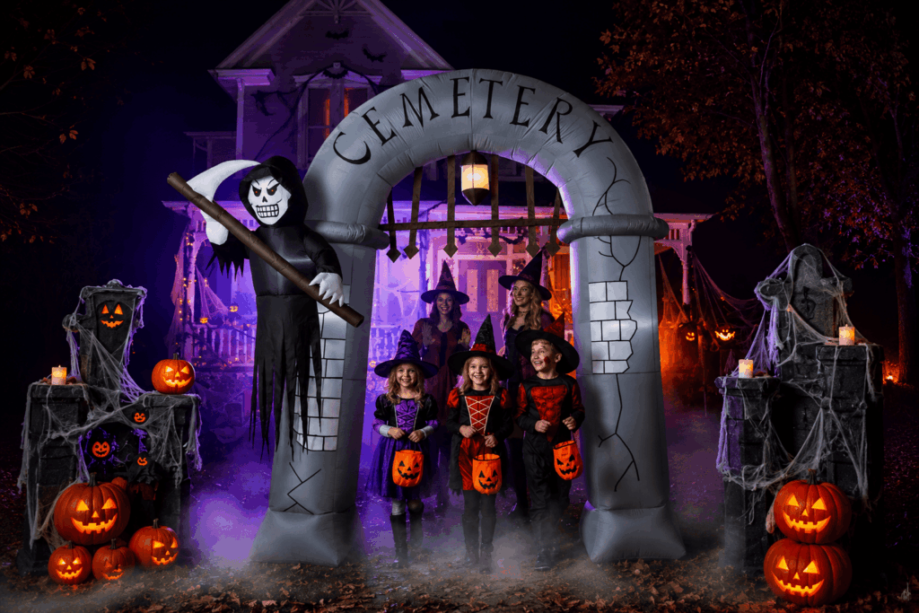 Une grande arche gonflable Halloween en forme d’entrée de cimetière est installée devant une maison décorée, avec enfants déguisés, citrouilles lumineuses, brume et éclairages violets et orange.