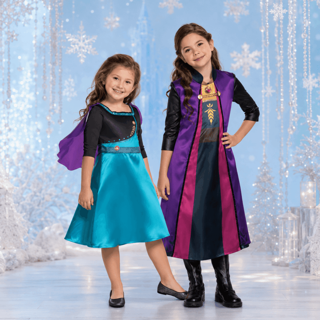 Deux fillettes posent côte à côte dans un décor bleu clair inspiré de l’hiver, avec flocons décoratifs et lumière douce. La plus petite porte une robe turquoise avec cape violette, la plus grande une robe violette et noire avec manteau assorti, dans une ambiance féerique idéale pour illustrer des déguisements princesse fille.